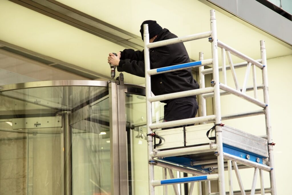 A maintenance worker on a scaffold is performing upkeep on a revolving door, showcasing the importance of regular maintenance to ensure the doors serve their purpose efficiently.